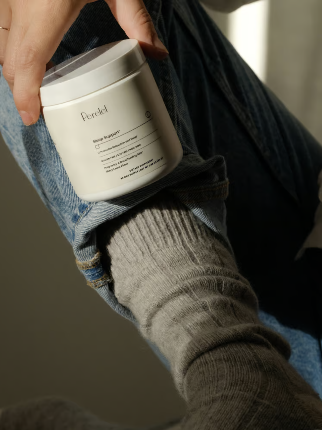 A person in jeans and gray socks is holding a white jar labeled "Perclel" with both hands.