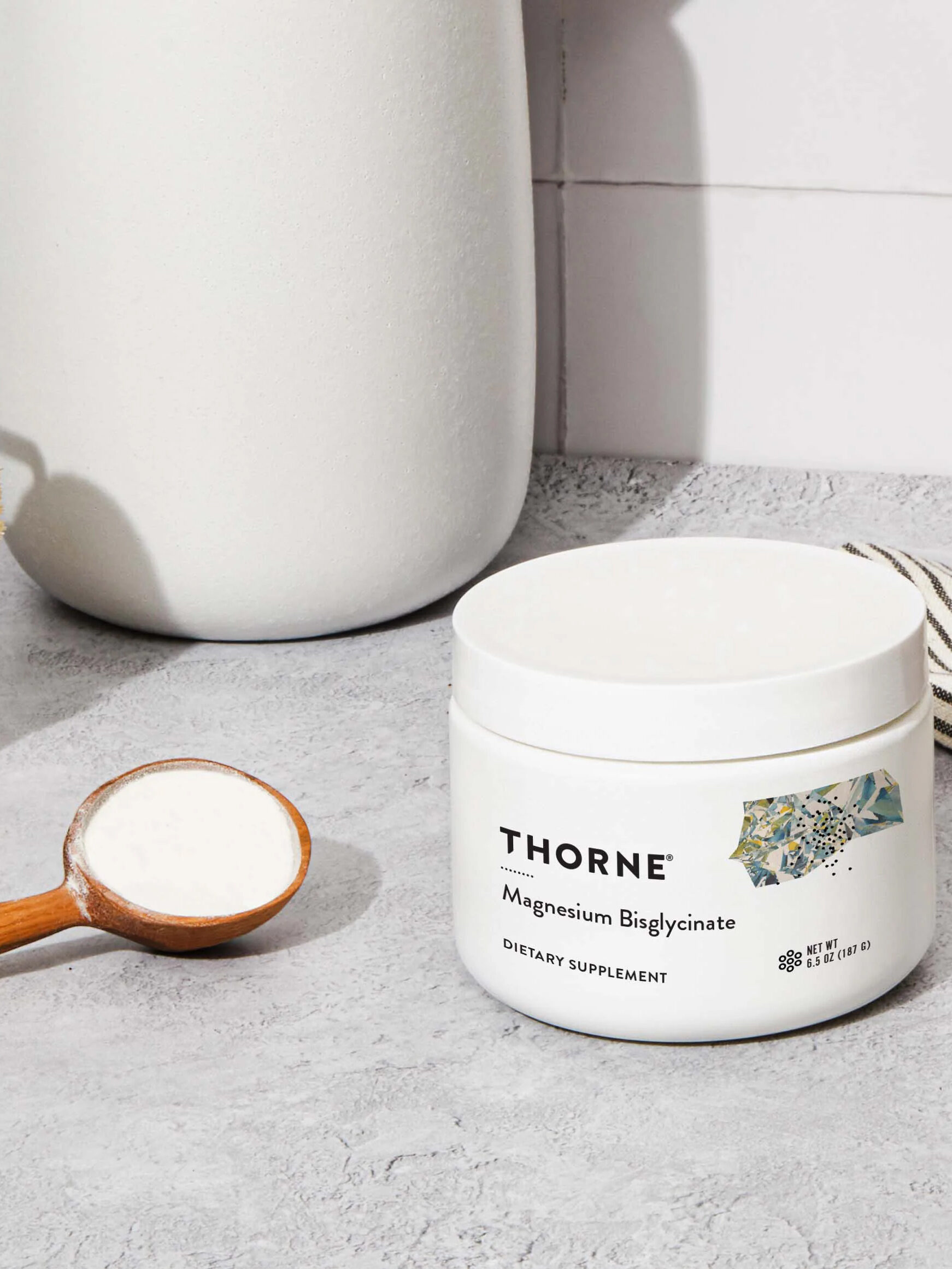 A jar of Thorne Magnesium Bisglycinate dietary supplement is on a countertop beside a wooden spoon filled with the powder.
