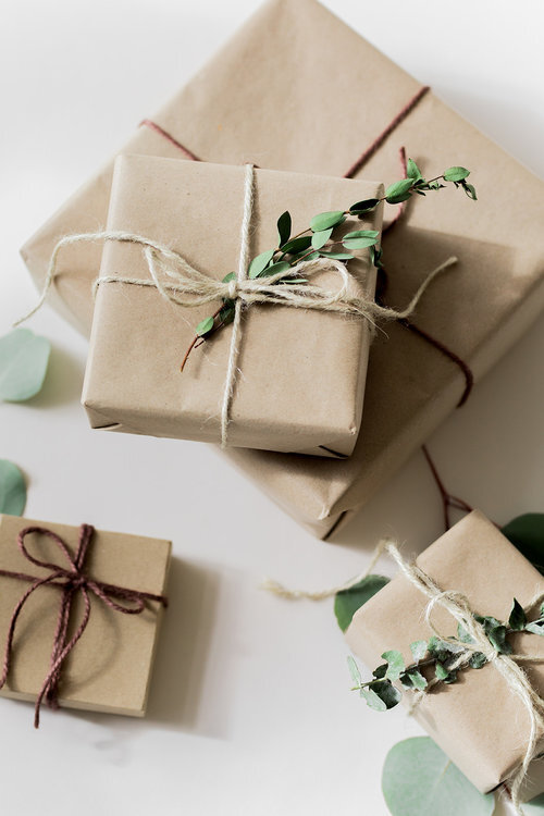 Three brown paper-wrapped gifts tied with twine and decorated with eucalyptus leaves are arranged on a white surface.