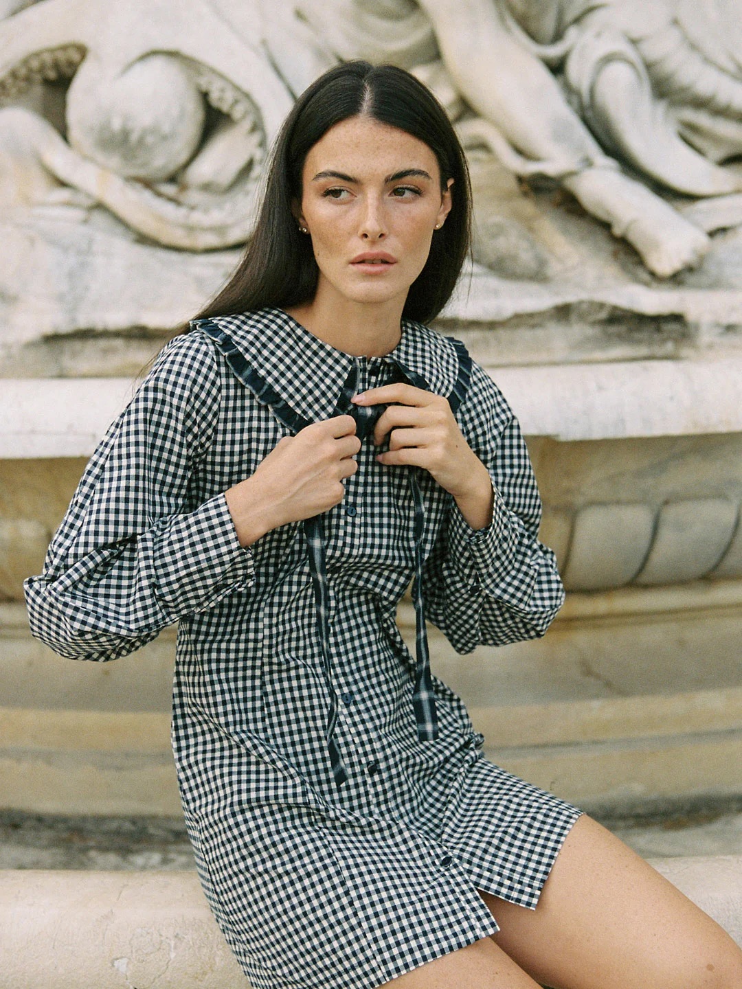 Woman with long dark hair wears a black-and-white checkered dress with a collar and ribbon, sitting in front of a stone fountain with sculpted details.