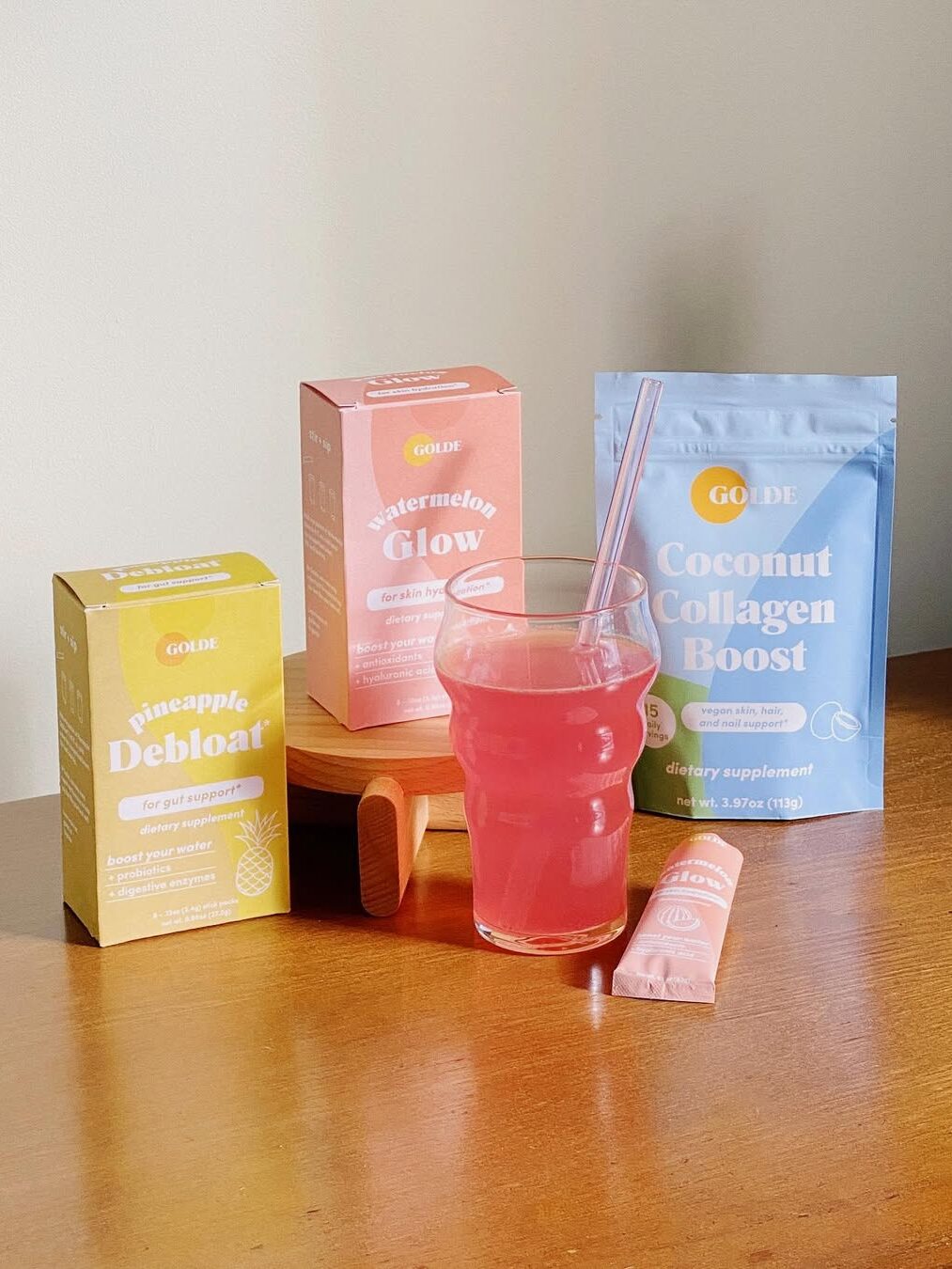 A glass of pink drink on a table with boxes labeled Pineapple Debloat, Watermelon Glow, and Coconut Collagen Boost, along with a small packet beside the glass.