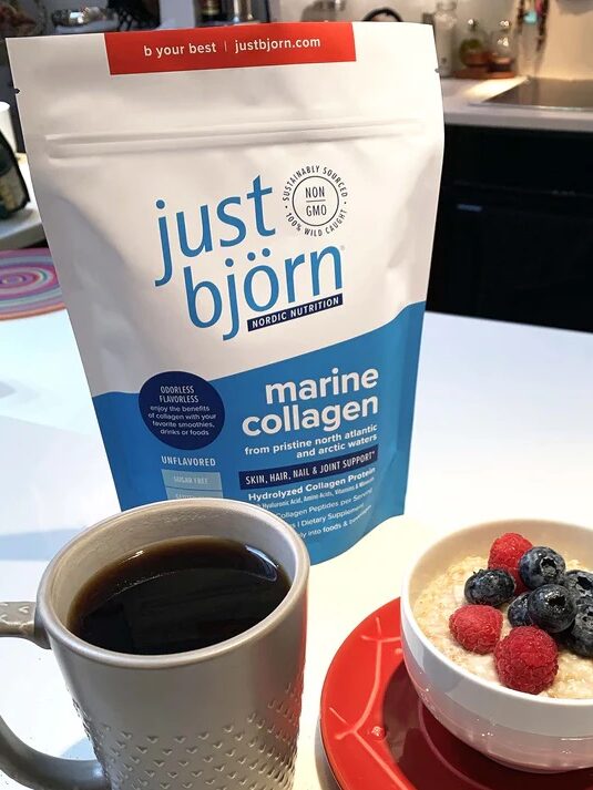 A cup of coffee, a bowl of oatmeal topped with berries, and a bag of Just Björn marine collagen on a kitchen counter.