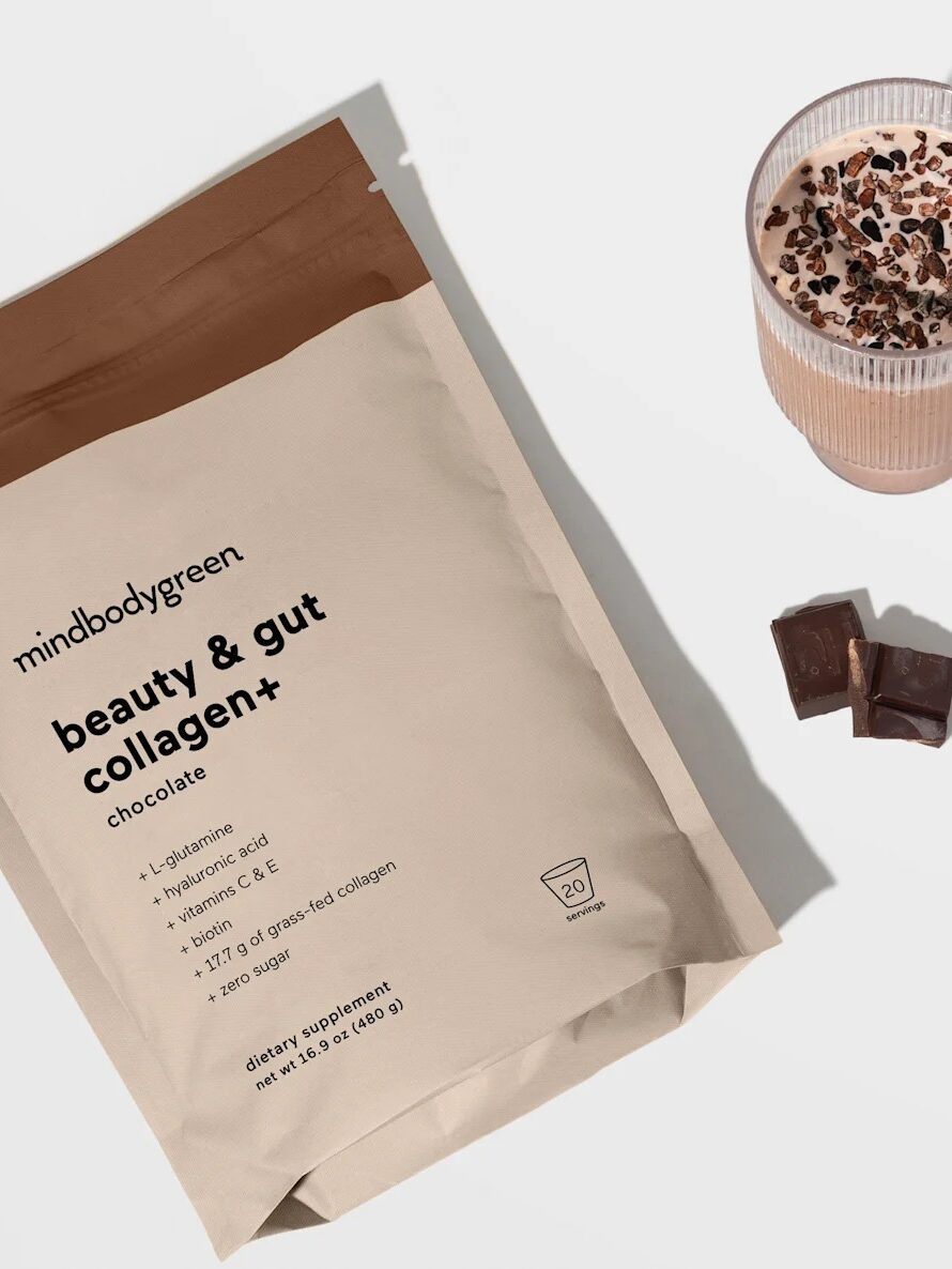 Bag of chocolate collagen powder next to a glass of smoothie topped with cacao nibs, two chocolate squares, and a golden spoon.