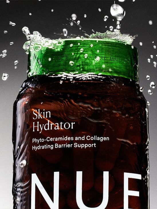 Close-up of a skincare bottle labeled "Skin Hydrator" with water splashing around it. The label mentions Phyto-Ceramides and Collagen for hydrating barrier support.