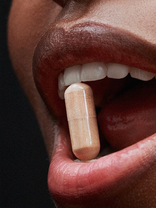 Close-up of a person holding a brown capsule pill between their teeth with their mouth open.
