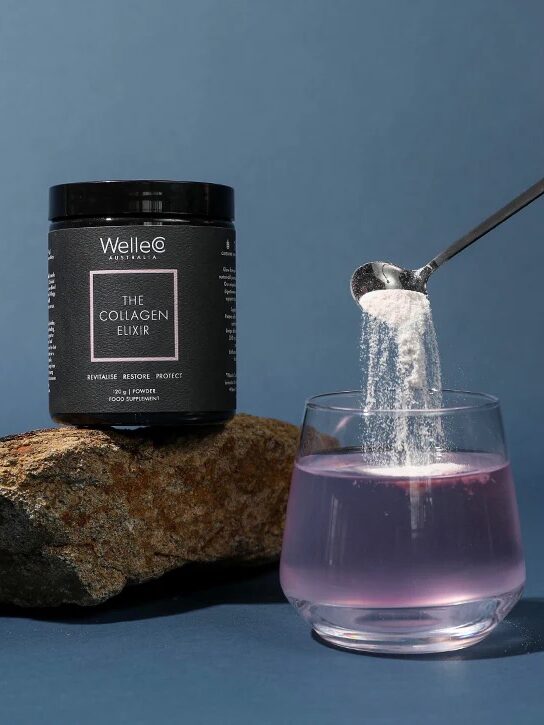 A hand pours collagen powder from a jar into a glass of pink liquid, with a rock in the background.