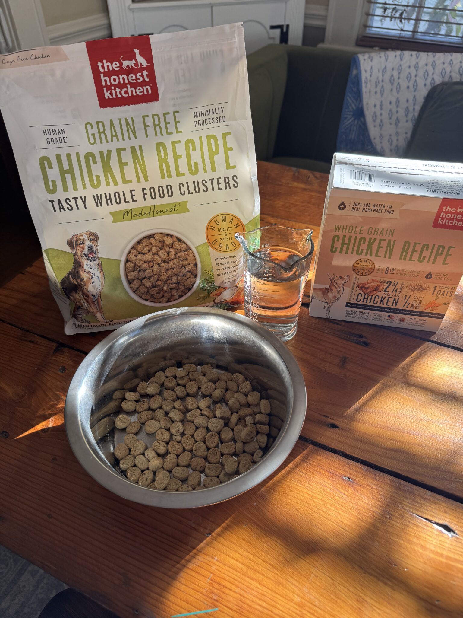 A bowl of dry dog food sits on a wooden table next to a glass of water and two packages of The Honest Kitchen dog food.