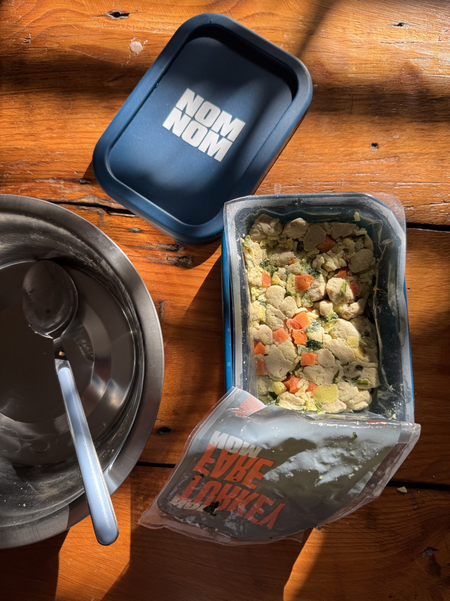 A container of Nom Nom pet food with chopped meat and vegetables beside an empty metal pet bowl and spoon on a wooden surface.