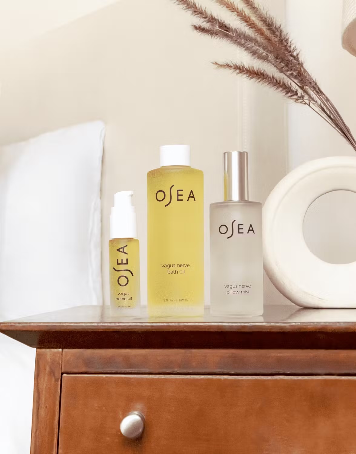 Three OSEA skincare products—a small pump bottle, a medium bottle, and a spray bottle—are displayed on a wooden nightstand next to a bed and a decorative vase with dried grasses.