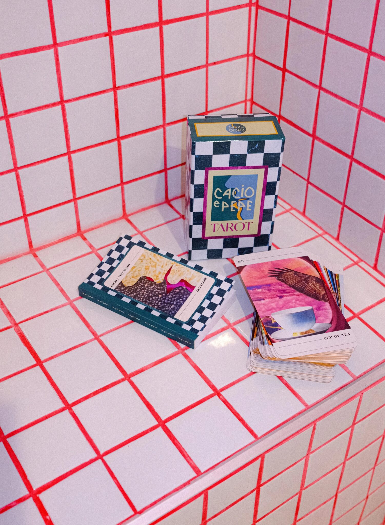 A tarot card deck and its box are placed on a tiled surface with red grid lines. Some cards are stacked face down while others are spread out showing colorful illustrations.