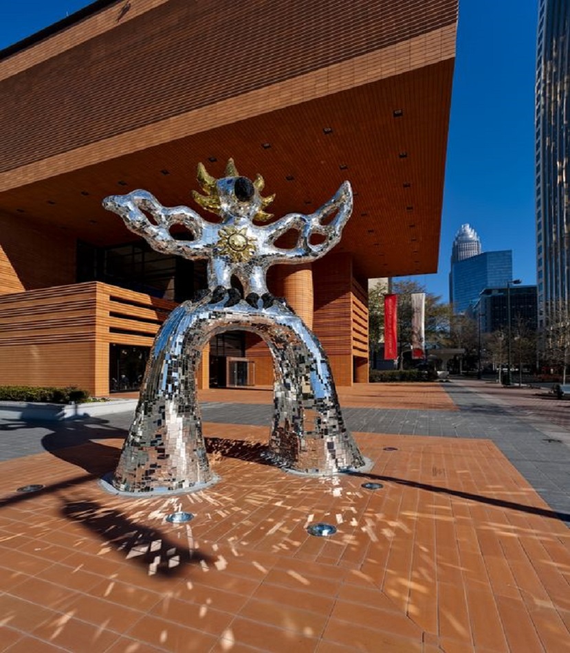 A large mirrored mosaic sculpture with abstract features stands outside a modern brick building in an urban setting on a sunny day.