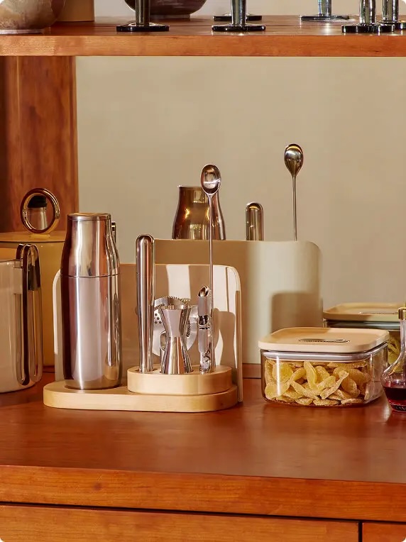 A wooden bar counter holding a cocktail shaker, various bar tools in a wooden holder, a glass container of dried fruit, and other drink accessories.