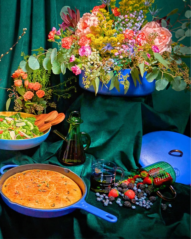 A vibrant spread featuring a salad, a baked dish in a blue pan, a glass oil bottle, assorted berries, and a floral arrangement against a green fabric backdrop.
