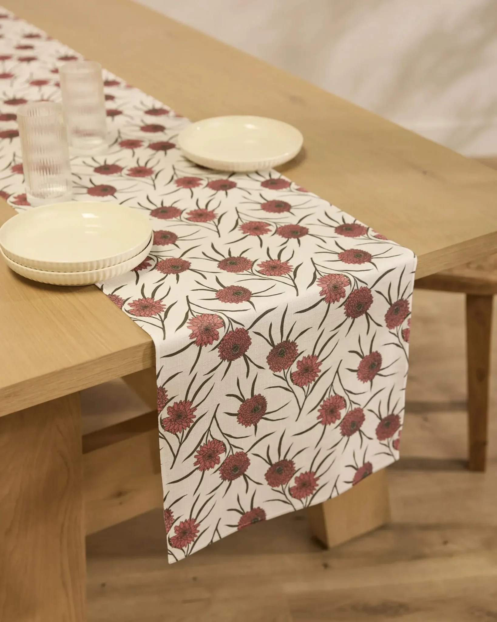 A wooden table set with cream plates and glasses, featuring a white table runner decorated with a red and green floral pattern.