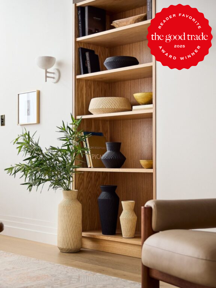 A wooden bookshelf with various decorative vases stands against a white wall. A potted plant is placed beside the shelf. A "The Good Trade 2025 Reader Favorite Award Winner" badge is visible.