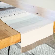 A wooden table with a neutral-toned, striped table runner draped over the edge, set on a light-colored woven rug.