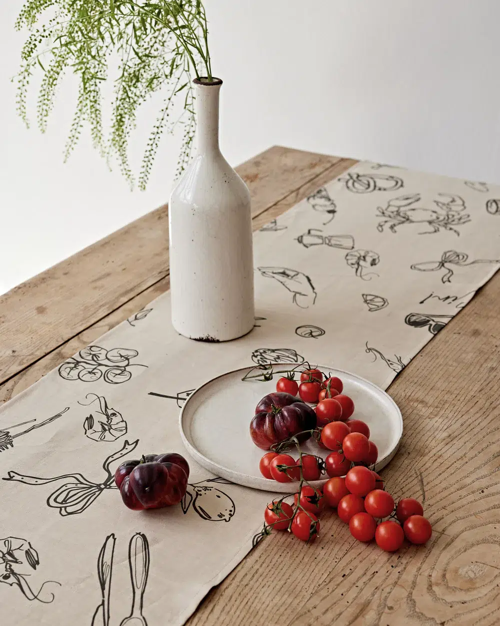 A white ceramic bottle with greenery, a plate with a dark tomato, and a cluster of cherry tomatoes on a rustic table with a patterned table runner.