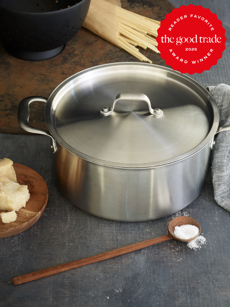 A stainless steel pot with a lid sits on a dark surface. Ingredients like cheese, spaghetti, and salt are nearby. A "The Good Trade 2025 Award Winner" badge is in the top right corner.