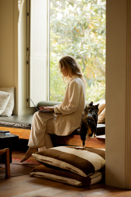 Person in a white outfit using a laptop by a window, with a cat walking nearby.