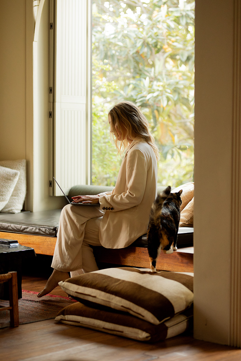 Person in a white outfit using a laptop by a window, with a cat walking nearby.