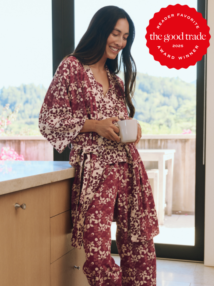 Woman in floral pajamas stands by a kitchen counter holding a mug, with a "The Good Trade" award badge in the corner.