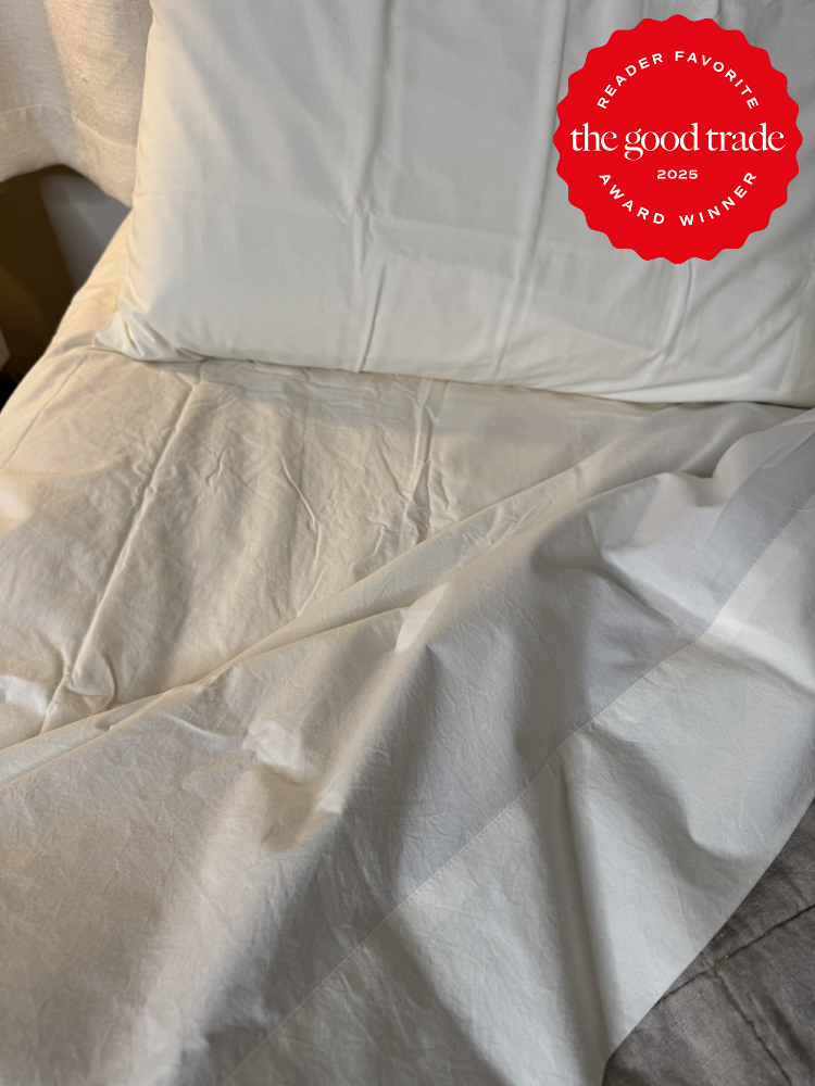 White bedding with a pillow on a bed. A red circular badge in the corner reads "Reader Favorite The Good Trade Award Winner 2025.