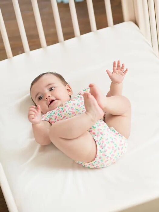 A baby in a patterned onesie lies on a white mattress in a crib, looking upward with arms raised and legs bent.