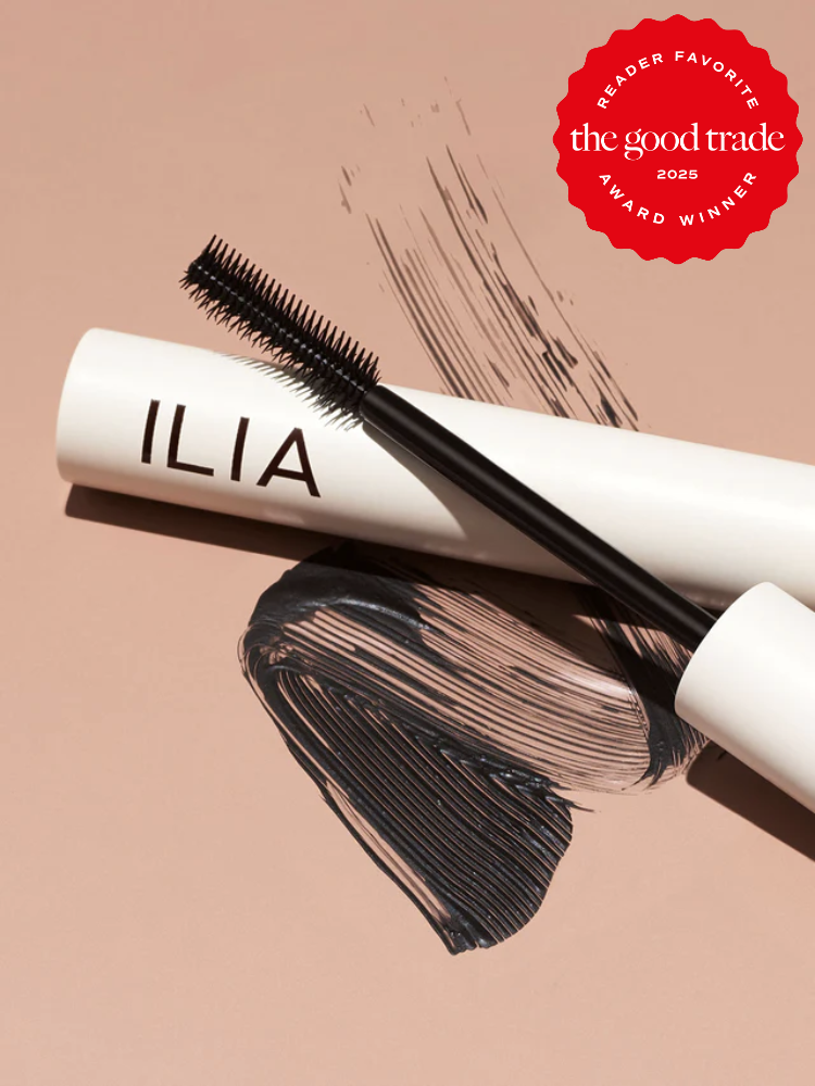 ILIA mascara on a tan surface, with a mascara wand and smudges of black mascara nearby. "The Good Trade 2025 Reader Favorite Award Winner" badge in the top right corner.