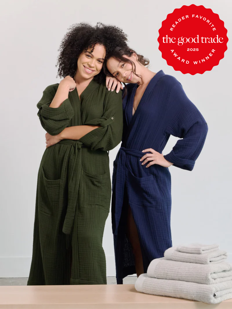 Two women wearing green and blue robes pose together. A red seal reads "Reader Favorite the good trade 2025 Award Winner." White towels are stacked on a wooden surface in front.