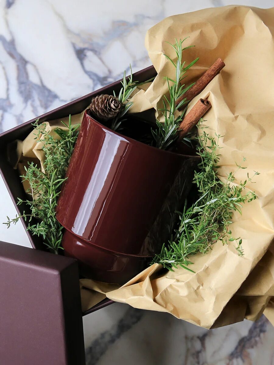 A brown ceramic mug filled with rosemary, cinnamon sticks, and pine cones sits on crumpled paper in an open box.