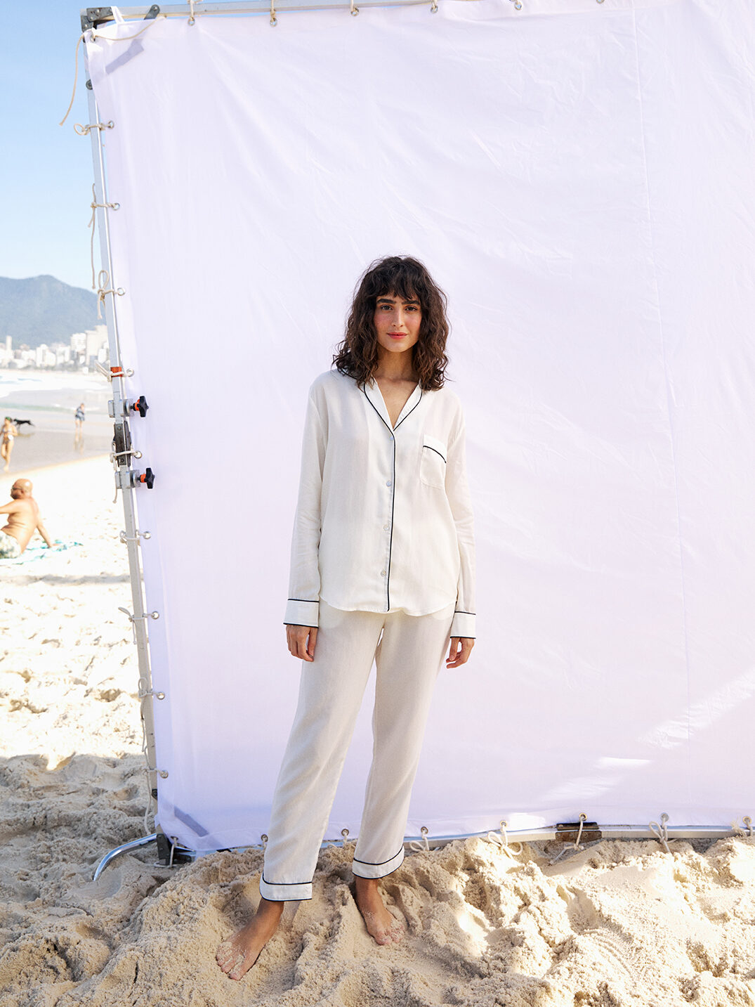 A model in front of a white photo backdrop on a beach wears white silk pajamas with black piping
