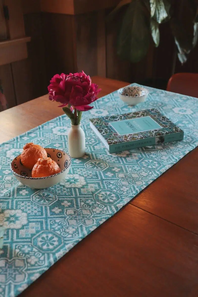 A wooden table with a blue patterned runner, a vase with red flowers, a bowl of oranges, and a book placed beside a small decorative dish.