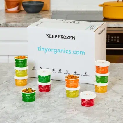 A box labeled "Keep Frozen" from tinyorganics.com on a kitchen counter with 12 assorted Tiny Organics food containers arranged in front of it.