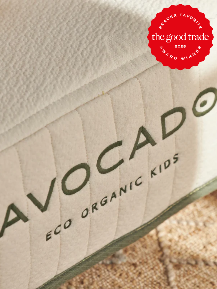 Close-up of a mattress with a label reading "Avocado Eco Organic Kids." The mattress has an off-white quilted surface, and a red award badge is visible in the top right corner.