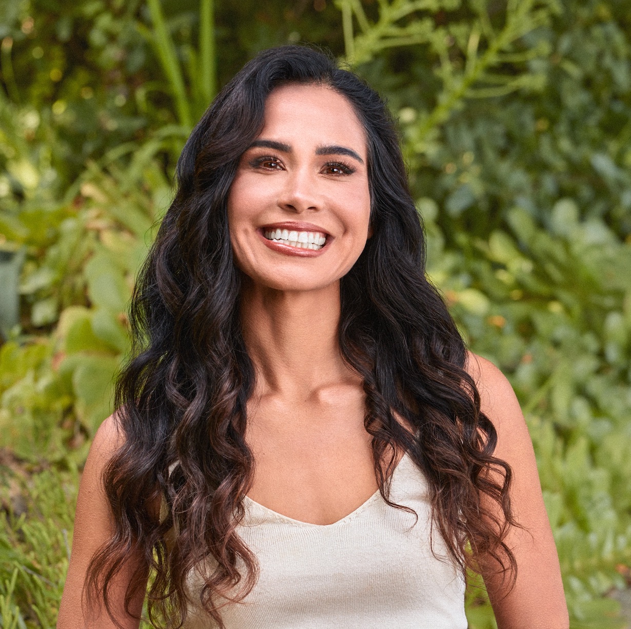 A person with long, dark, wavy hair smiles broadly in front of a background of green foliage.