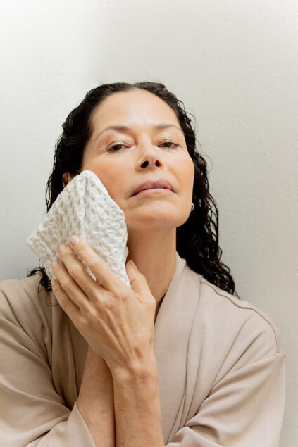 Person with wet hair gently pressing a textured cloth to their cheek, wearing a beige robe, against a pale background.