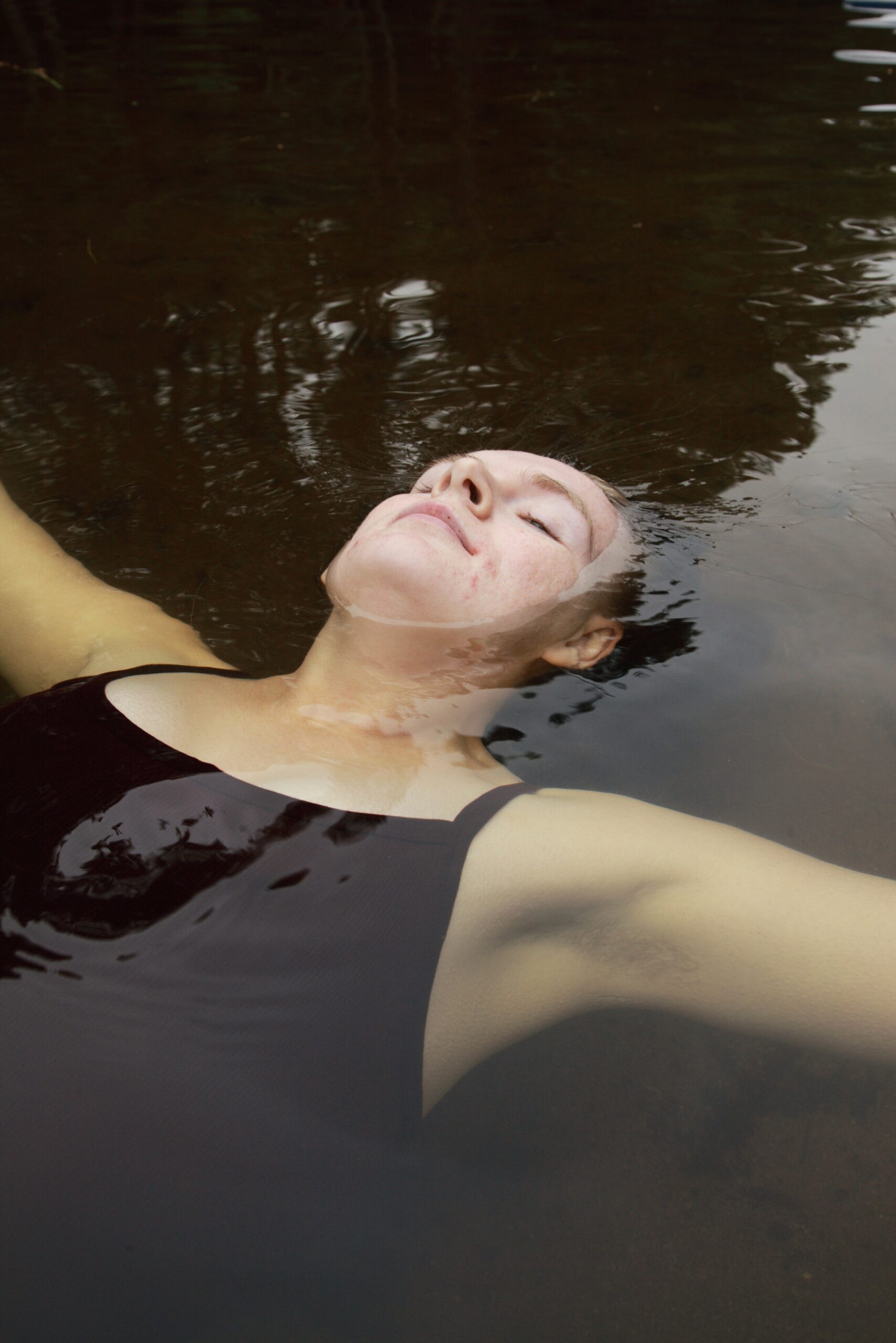 Person in a black tank top floating calmly on their back in dark water with eyes closed.