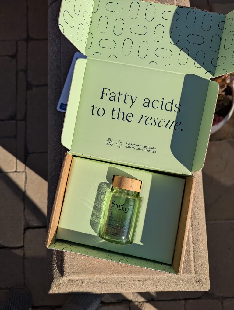 Open cardboard box with a small glass bottle inside. The lid is wooden, and the box lid reads, "Fatty acids to the rescue.