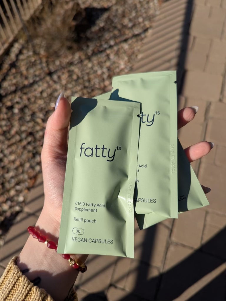 Person holding two green packets labeled "fatty15" with "C15:0 Fatty Acid Supplement" and "VEGAN CAPSULES" printed on them. Stone path and plants visible in the background.