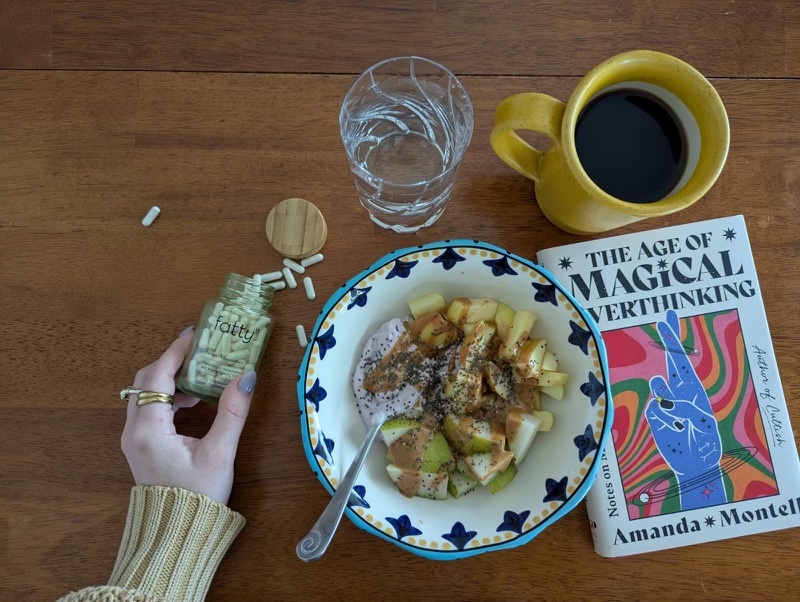 Breakfast with a bowl of yogurt, apple chunks, and chia seeds, a glass of water, cup of coffee, a book titled "The Age of Magical Verthinking," pills, and a hand holding a bottle labeled "fatty.