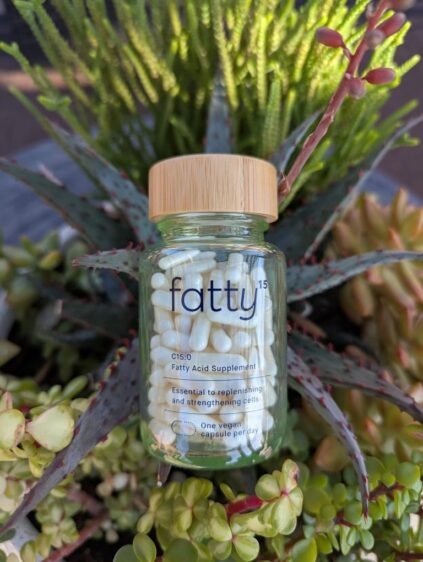 A bottle of fatty acid supplement capsules labeled "fatty" is placed in front of a background of mixed greenery and succulents. The bottle has a cylindrical wooden lid.