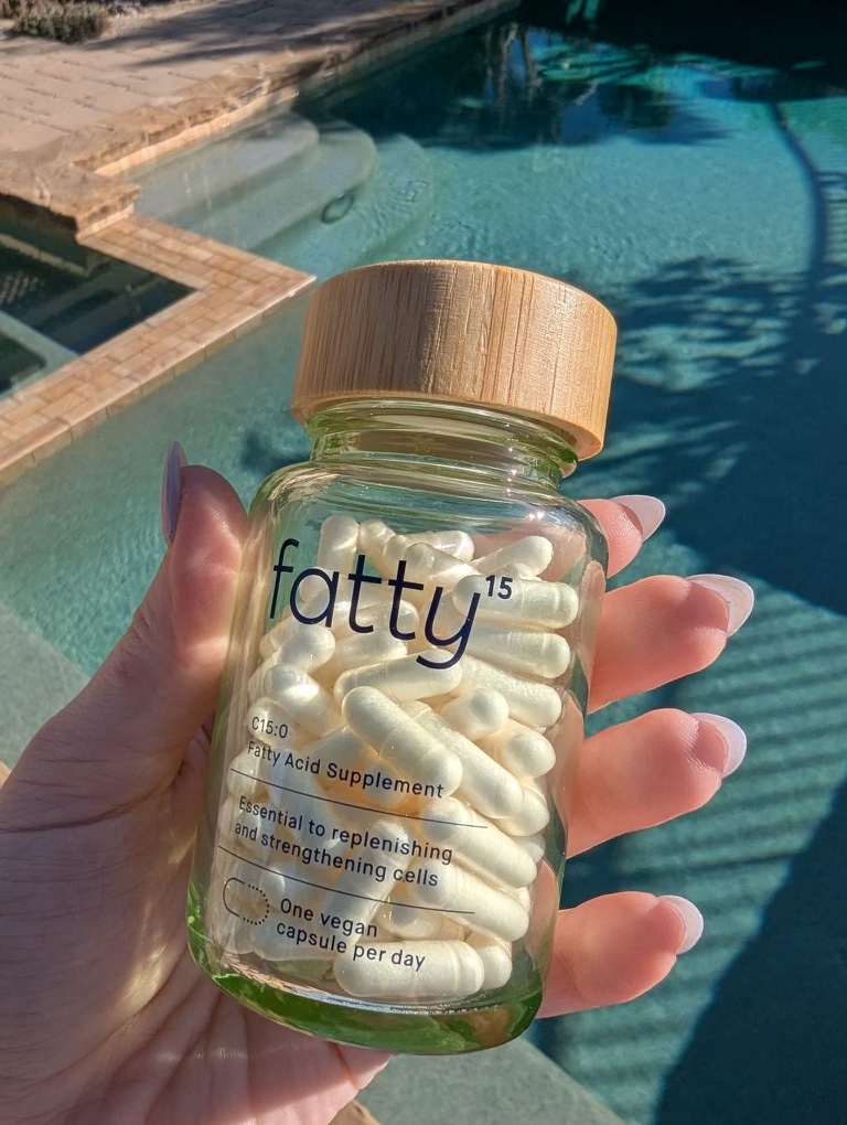 A hand holds a glass jar labeled "fatty15," containing fatty acid supplement capsules, by a swimming pool.