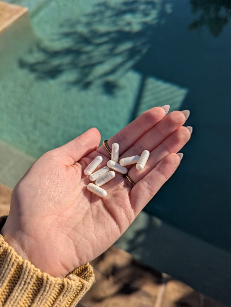 A hand holding white capsules, with a pool and patio in the background.