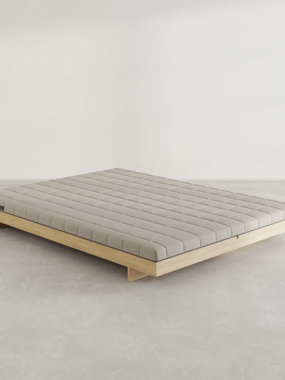 A minimalist bed with a light wood frame and a grid-patterned mattress on a plain floor, against a white wall.