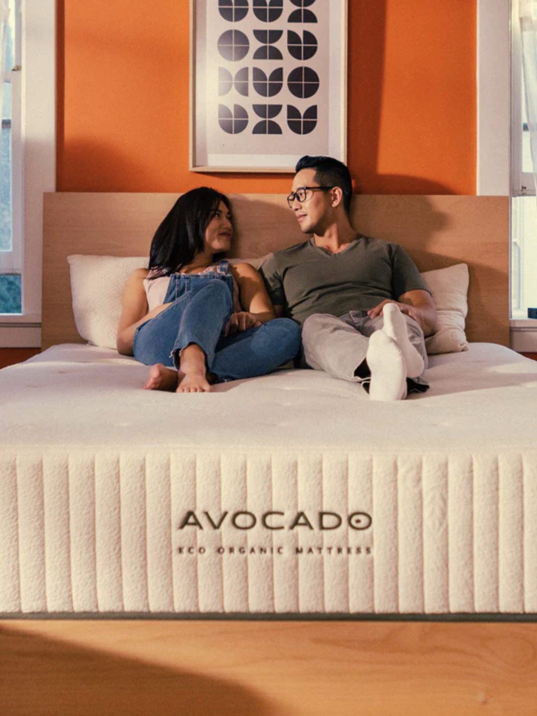 A couple sits on an Avocado eco organic mattress in a bedroom with orange walls and patterned artwork above the bed.