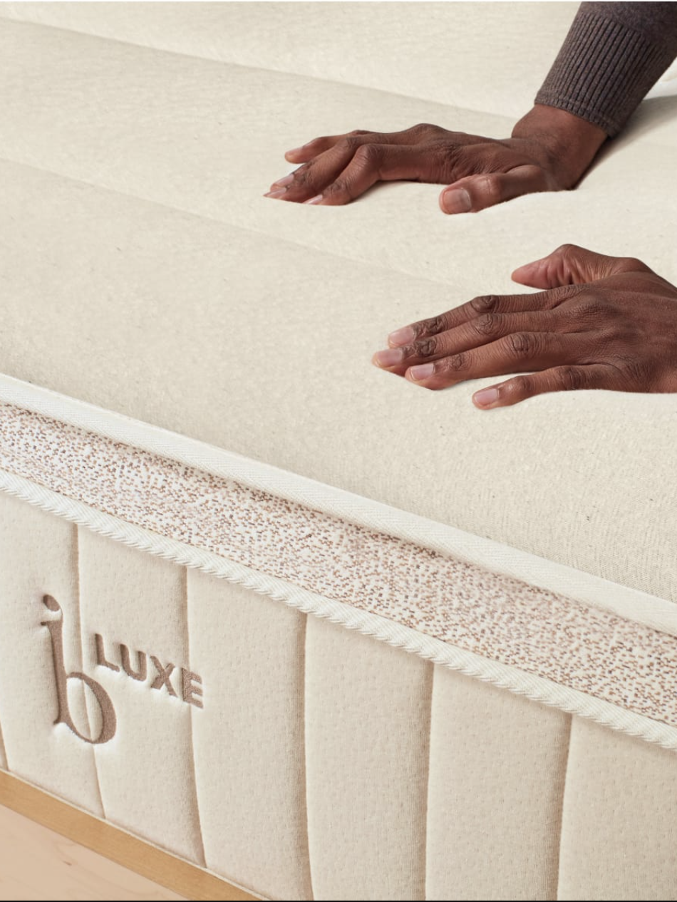 Two hands pressing down on a white mattress with "LUXE" embroidered on the side, demonstrating its softness and texture.