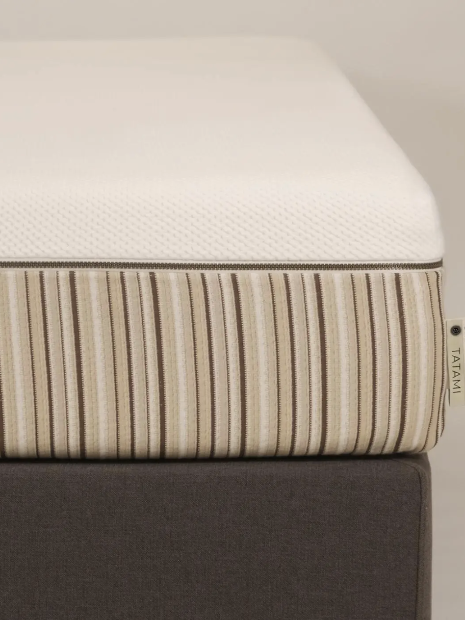 A close-up of a striped mattress topper on a dark bed base, with a cream tag labeled "ESSENTI TATAMI" attached to the side.