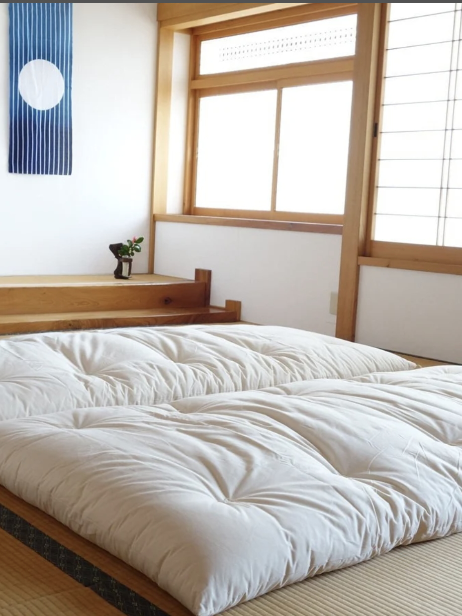 A traditional Japanese room features tatami mats and two white futons. A window and wall hanging with a circular design are visible in the background.