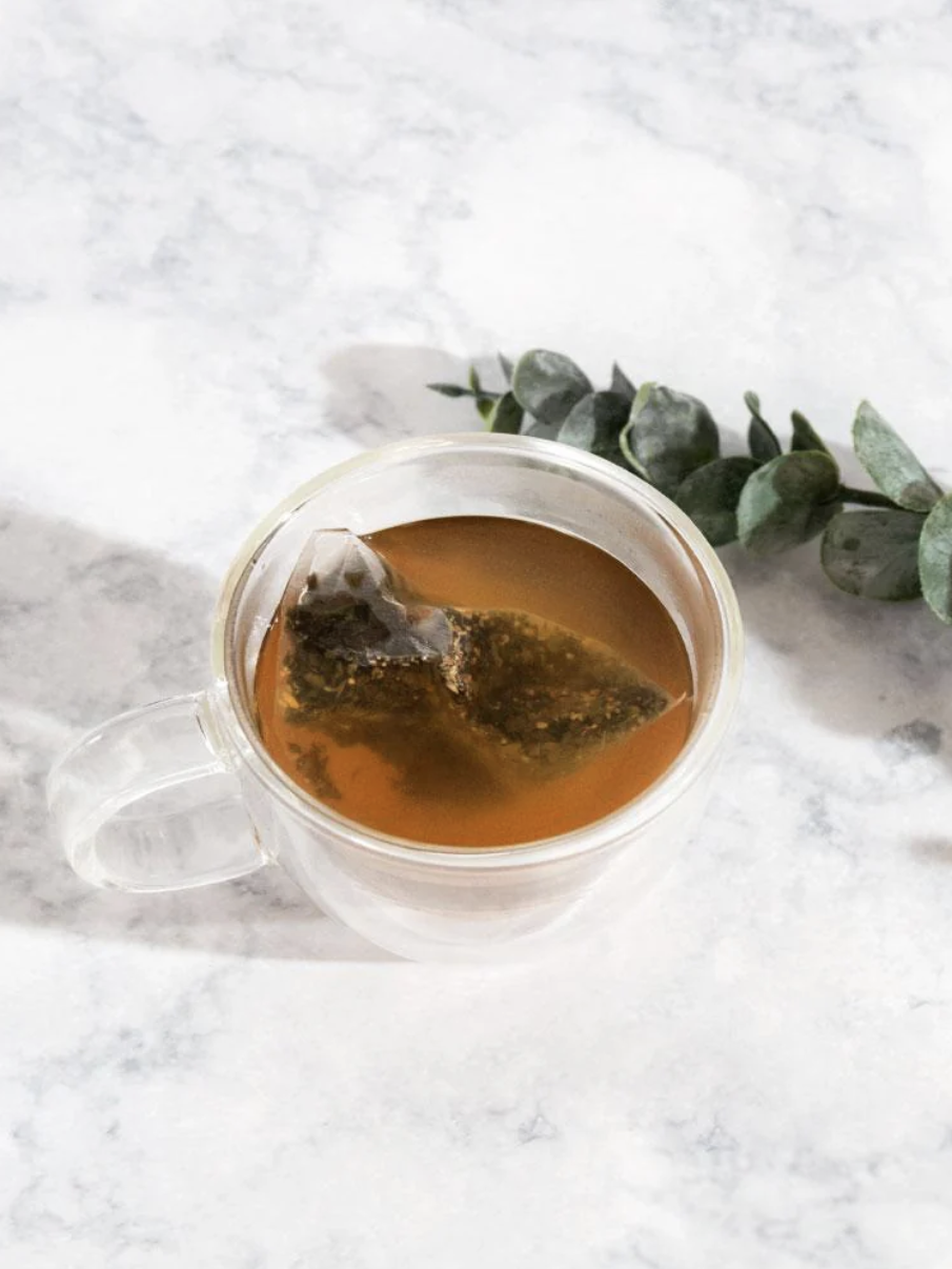 A clear glass cup with a tea bag inside sits on a marble surface next to a small branch of eucalyptus leaves.