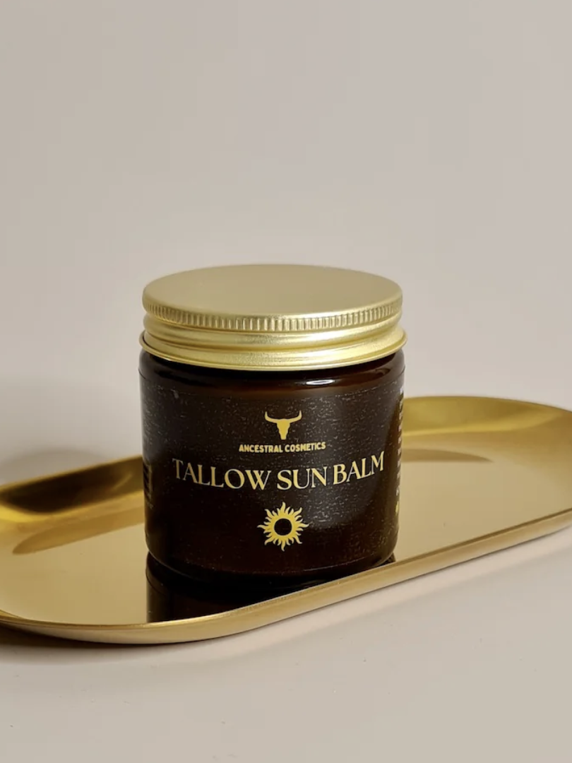 A jar of tallow sun balm with a gold lid sits on an oval, gold-colored tray against a plain background.